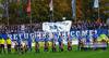 Fanprojekt Oldenburg gegen Rassismus mit Banner im Stadion hinter Fußballmannschaft mit Refugees Welcome